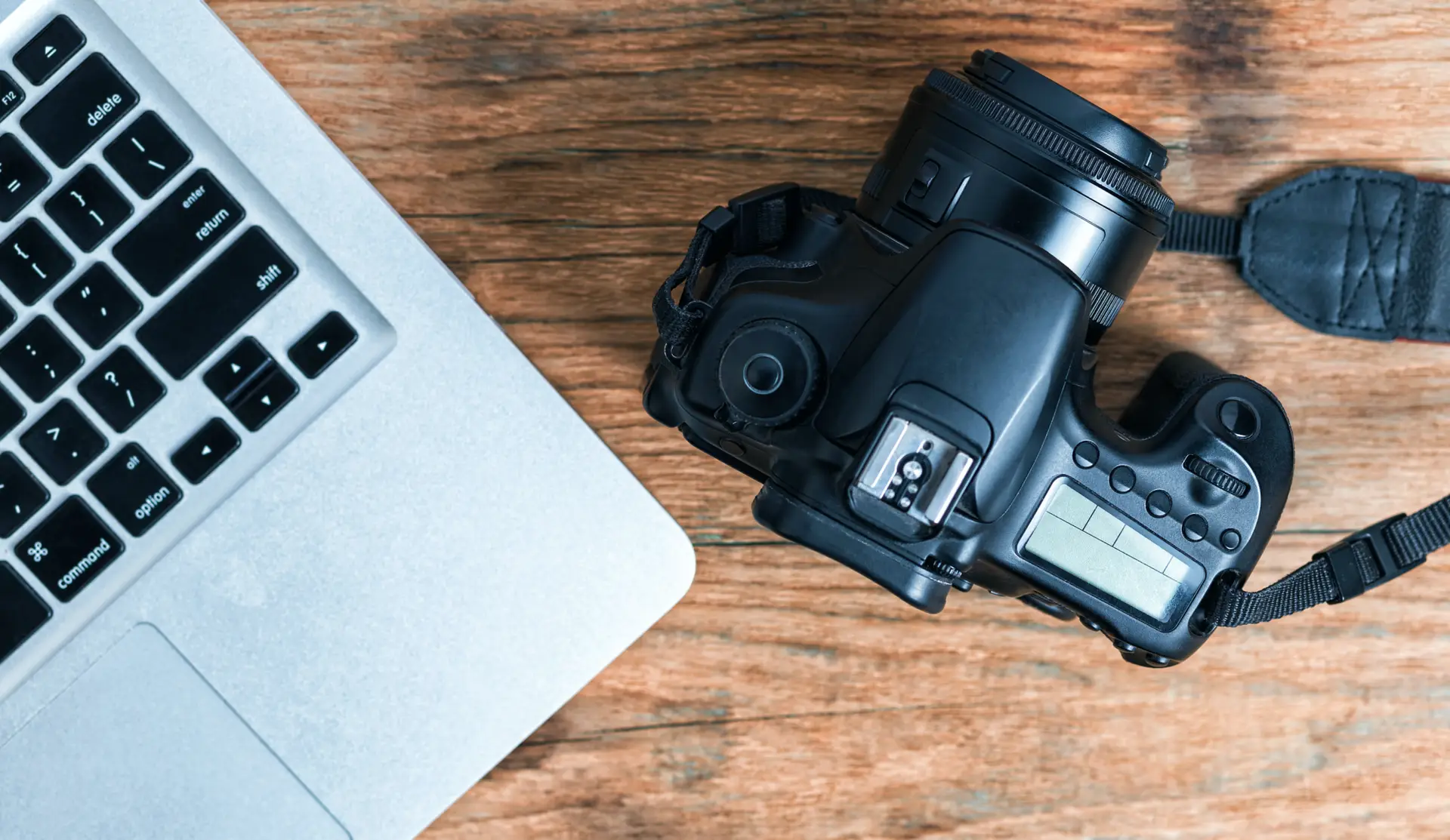 A camera rests next to a laptop on top of a wooden desk.