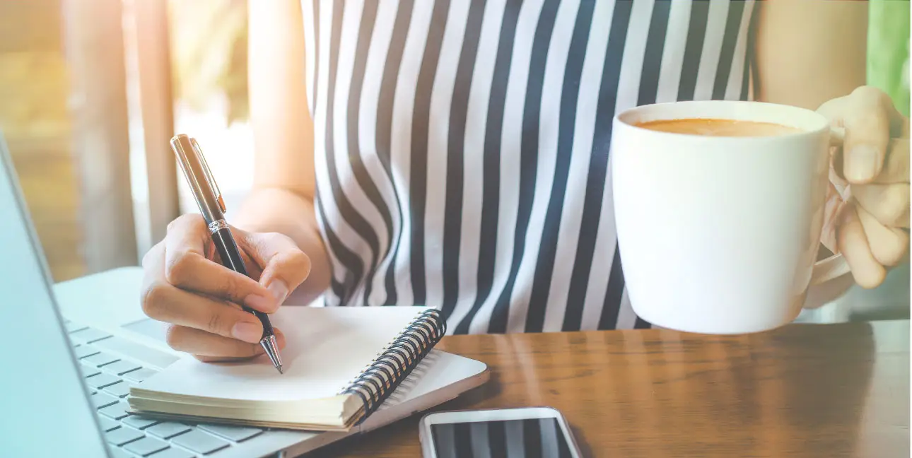A woman writes on a notepad that's resting on top of her laptop while she drinks coffee.