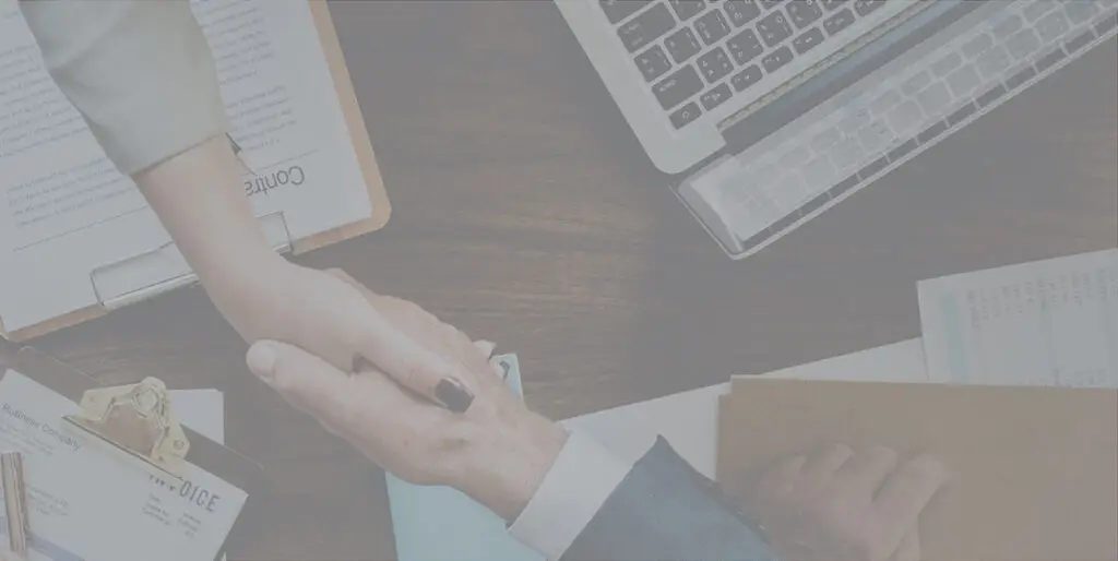 Two people shake hands over a conference table littered with laptops and papers