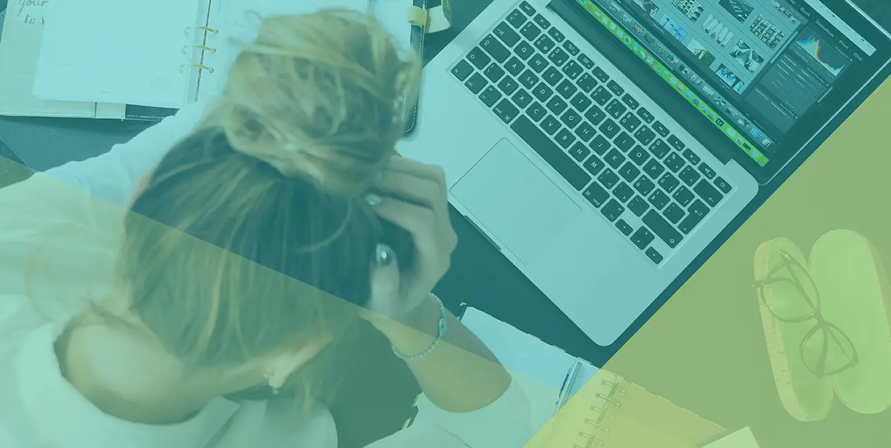 An employee holds their head in distress while surrounded by a computer and other work materials