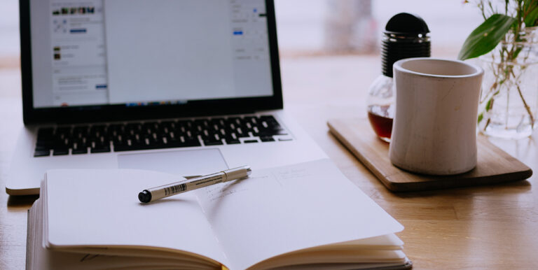 A laptop sits open while a Micron pen rests on a notebook filled with sketches.