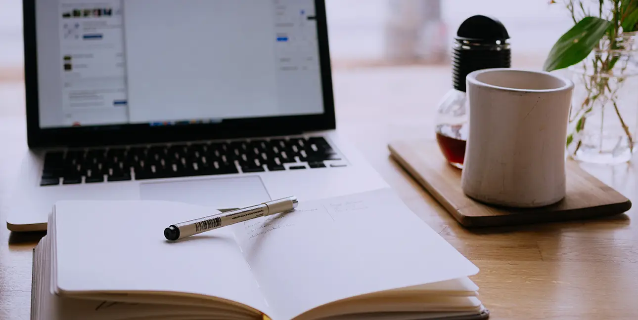 A laptop sits open while a Micron pen rests on a notebook filled with sketches.