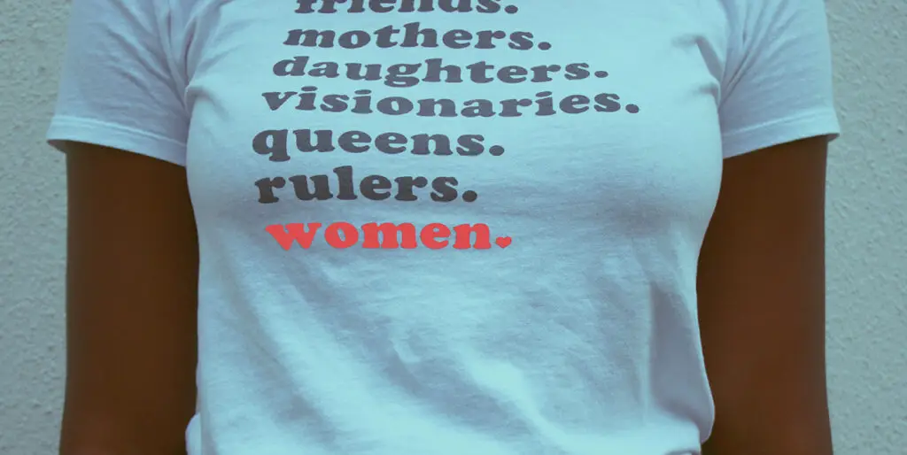 A woman wears a shirt that reads, "Friends. Mothers. Daughters. Visionaries. Queens. Rulers. Women."