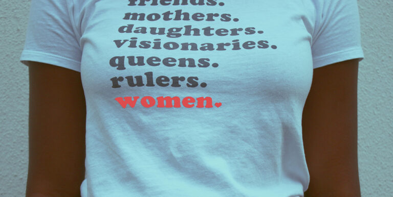 A woman wears a shirt that reads, "Friends. Mothers. Daughters. Visionaries. Queens. Rulers. Women."