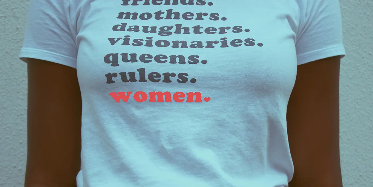 A woman wears a shirt that reads, "Friends. Mothers. Daughters. Visionaries. Queens. Rulers. Women."