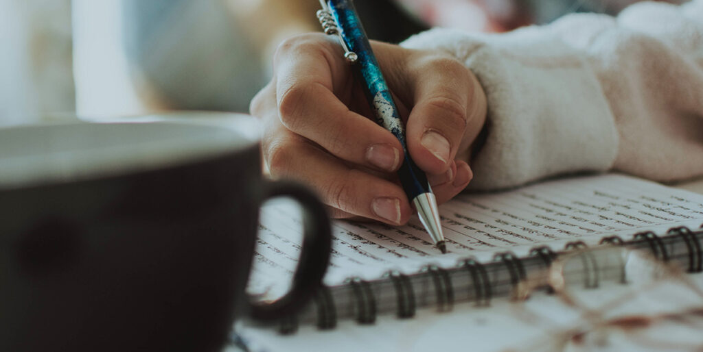A woman writes in a spiral notebook with a a coffee mug next to her.