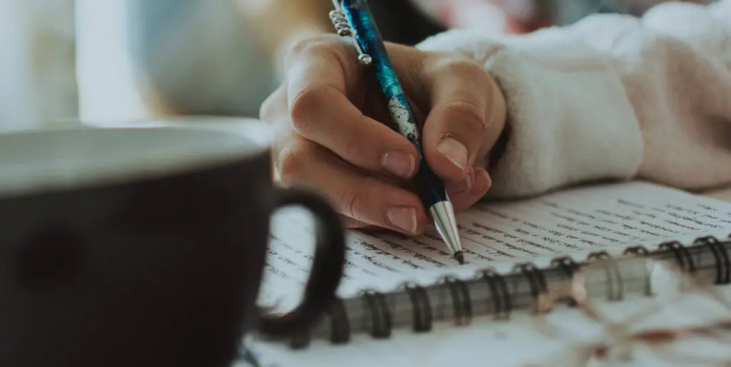 A woman writes in a spiral notebook with a a coffee mug next to her.