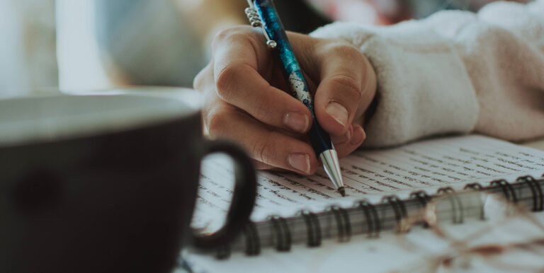 A woman writes in a spiral notebook with a a coffee mug next to her.