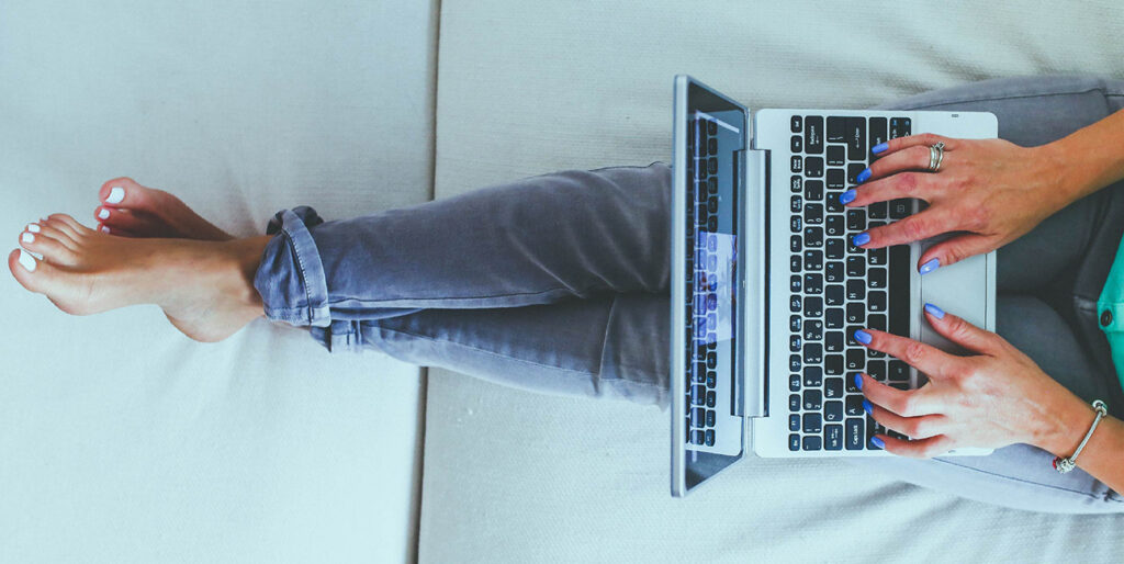 An image of a girl sitting on a bed, wearing jeans, typing on a laptop.