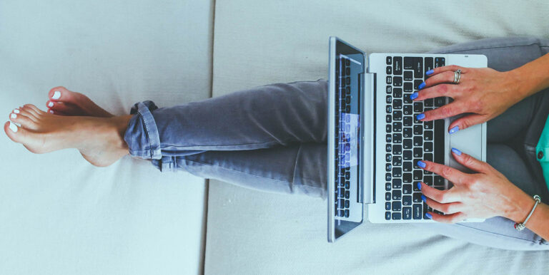 An image of a girl sitting on a bed, wearing jeans, typing on a laptop.
