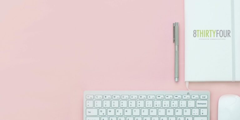 An image of a laptop, pen and journal against a pink table.