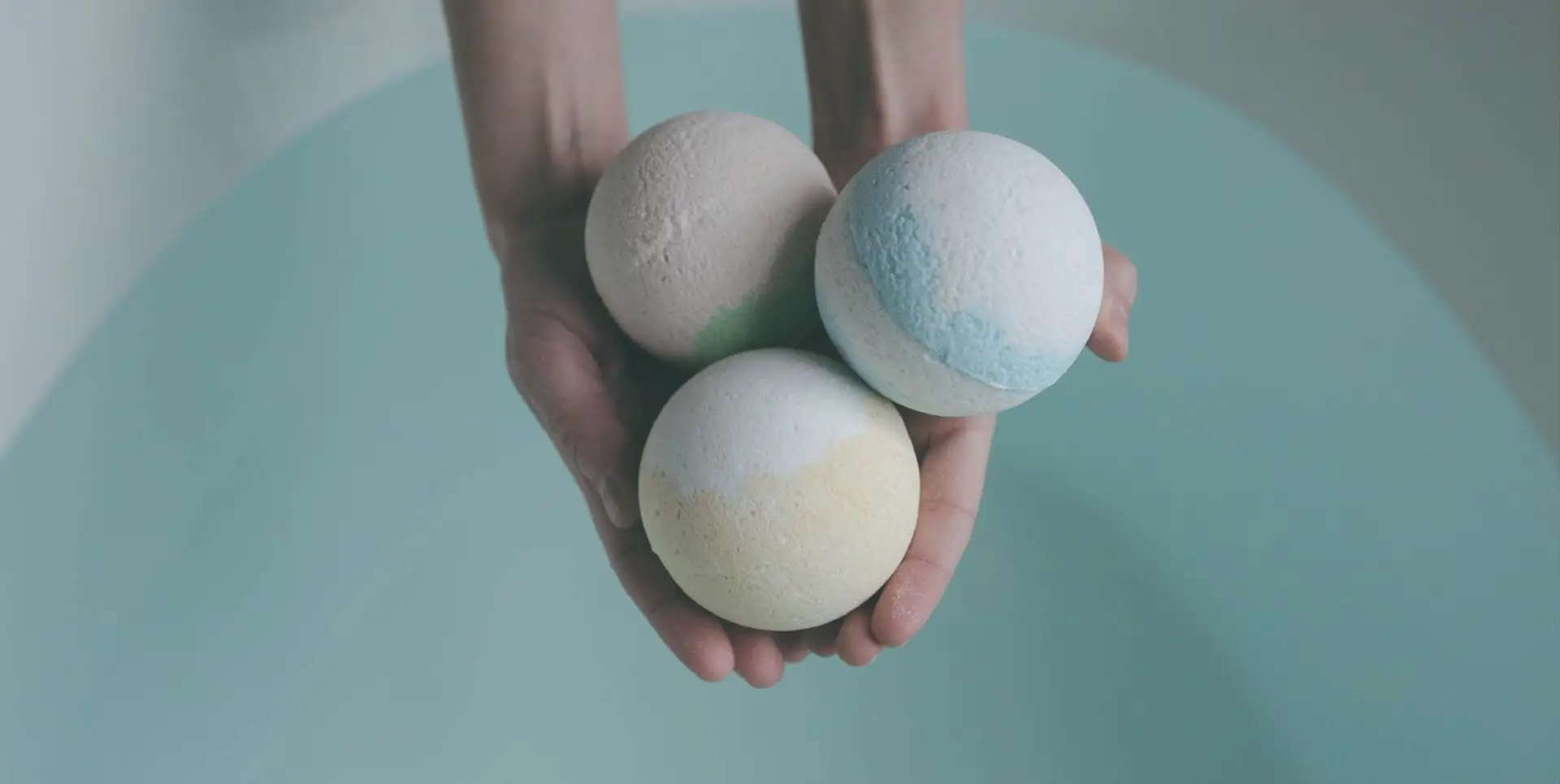 A person holding three bath bombs over a full tub of water.