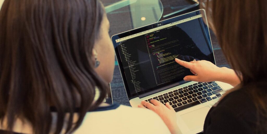 Two women looking at a computer screen with website code displayed on the screen. One of the women is pointing at the screen.