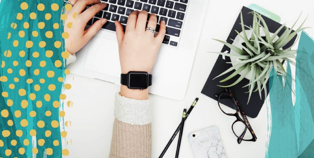 A person wearing a cuff bracelet and a watch, typing on a laptop. A pad of paper with a plant on top, and some eyeglasses and pens, lay next to it.