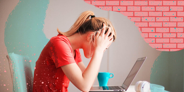 A woman sits at a desk with a cup of coffee and her laptop, her head in her hands.