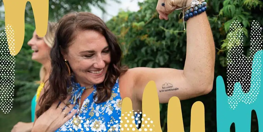A woman smiling and flexing her arm to show off a tattoo that says "find comfort in the uncomfortable."