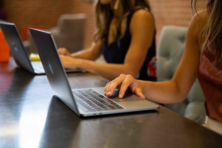 Two girls sitting at a desk typing on laptops.