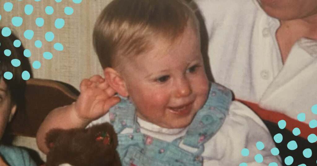 A blonde baby wearing blue overalls smiles at the camera from an aunt's lap.