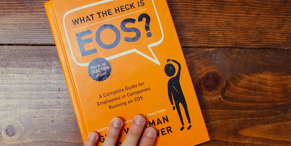 What the Heck is EOS book is laying on a wood table with a hand placed on the book. The background of the book is organge with black and white lettering.