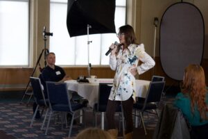 Kim stands at the front of a room. She's holding a microphone giving a speech to a room full of PR professionals.