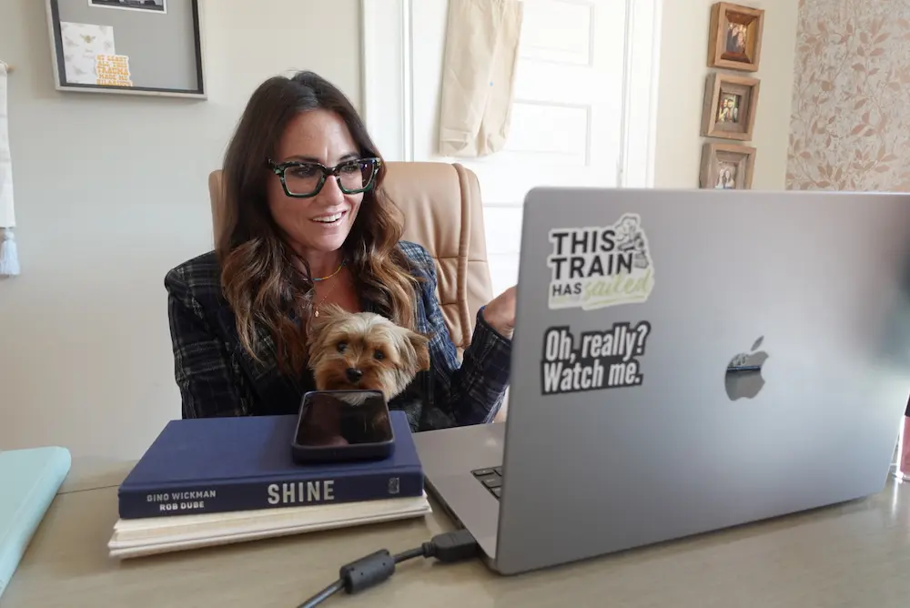 Kim Bode sits at her desk, laptop open while holding her dog. She smiles as she's on call.