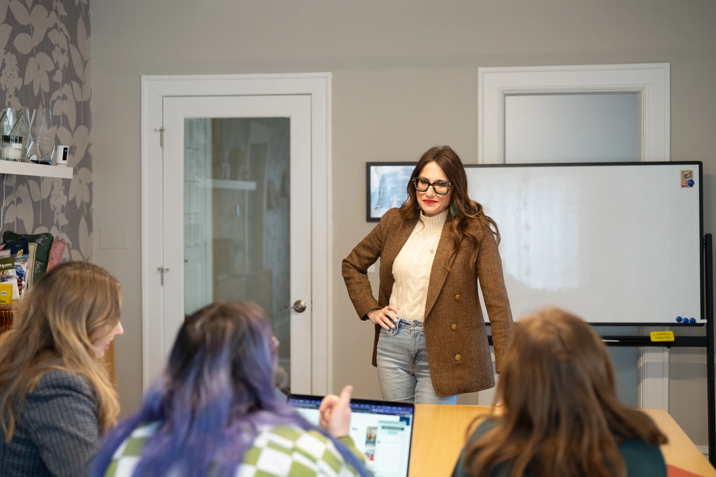 Kim stands in front of 3 women, her hand is on her hip and she's in front of a whiteboard smiling.